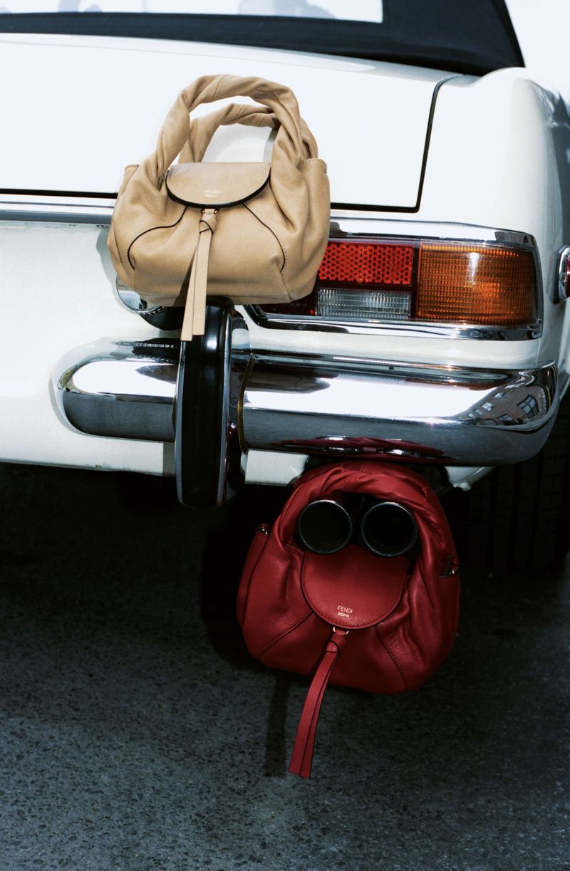 Fashion shot featuring a white vintage car with two handbags attached to the bumper and exhaust pipes, one tan and one red.