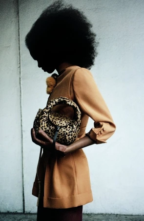 A person with an afro hairstyle wearing an orange blazer holds a leopard print handbag in front of a neutral wall background.