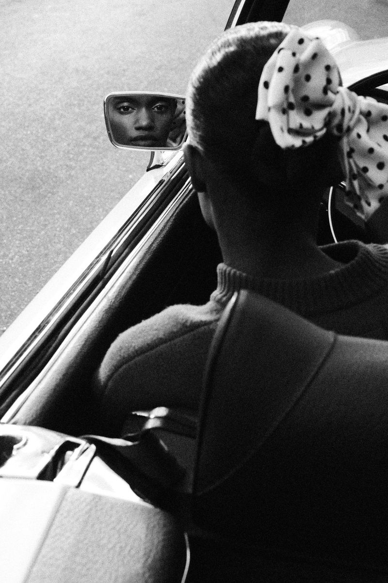 Black-and-white shot inside a car with an adult female glancing into the rearview mirror, her face reflecting in the mirror.