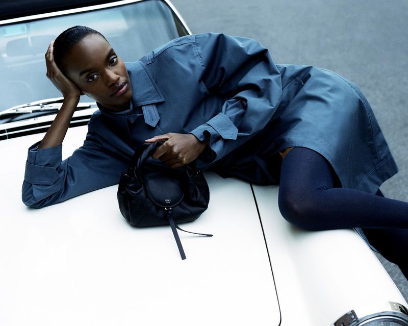 Fashion shot of an African-American woman in a trench coat lying on a white car hood with a black purse in her hand.
