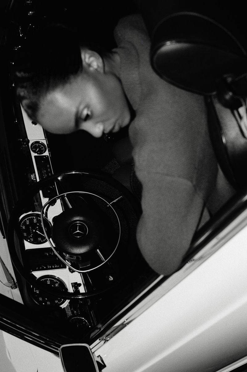 Monochrome portrait of an adult woman inside a classic car, focusing on her face, the steering wheel, and dashboard details.