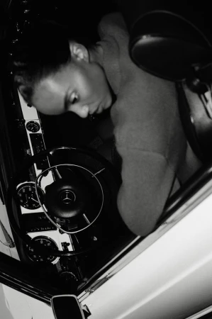 Monochrome portrait of an adult woman inside a classic car, focusing on her face, the steering wheel, and dashboard details.