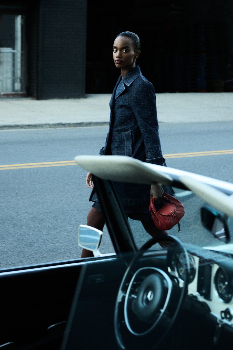 Woman in coat crossing street near classic convertible car, holding red leather purse on city street, dark mood, fashion editorial.