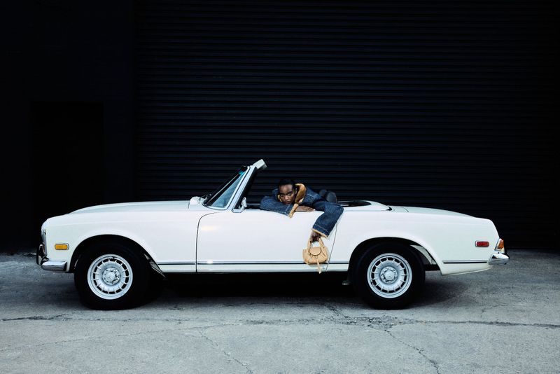 Model Farhiya Shire leaning out of a vintage white convertible car holding a small tan handbag against a dark backdrop.