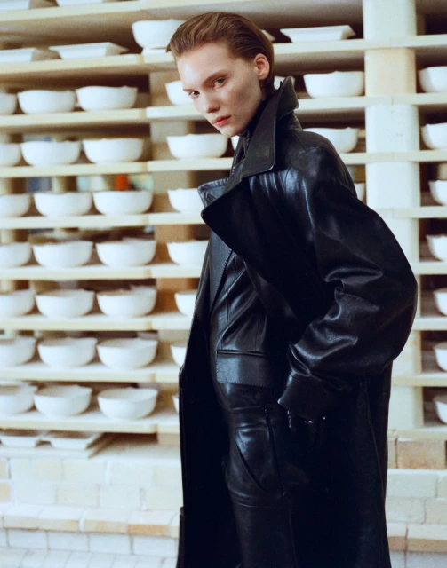 Fashion shot of a fair-skinned woman wearing a long black leather coat and pants, standing indoors in front of shelving with white bowls.