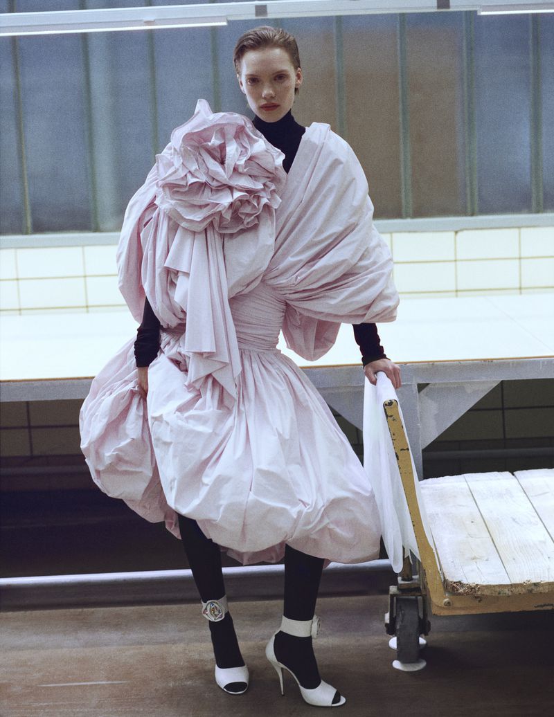 Elegant woman in a voluminous pink dress and black leggings poses with a metal cart in an industrial interior, creating a fashion editorial shot.