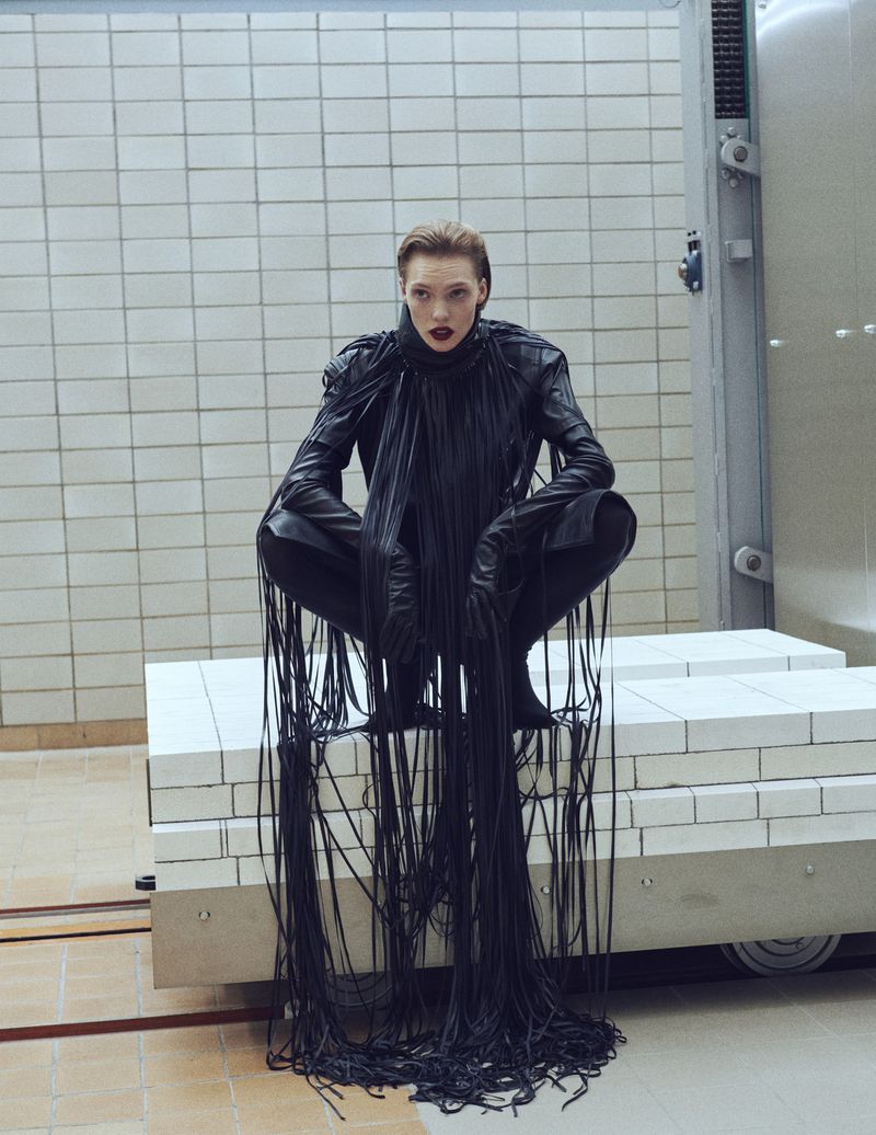 A woman in a black fringe outfit crouches on a white platform in a room with tiled walls. Bold red lipstick complements the black outfit.