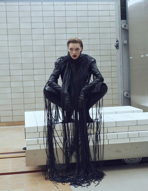 A woman in a black fringe outfit crouches on a white platform in a room with tiled walls. Bold red lipstick complements the black outfit.