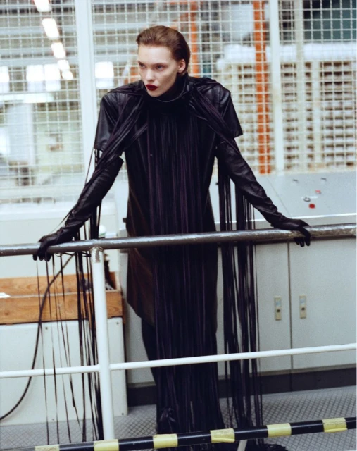 Fashionable adult woman wearing long black dress and gloves in an industrial location with white and orange machinery in the background.