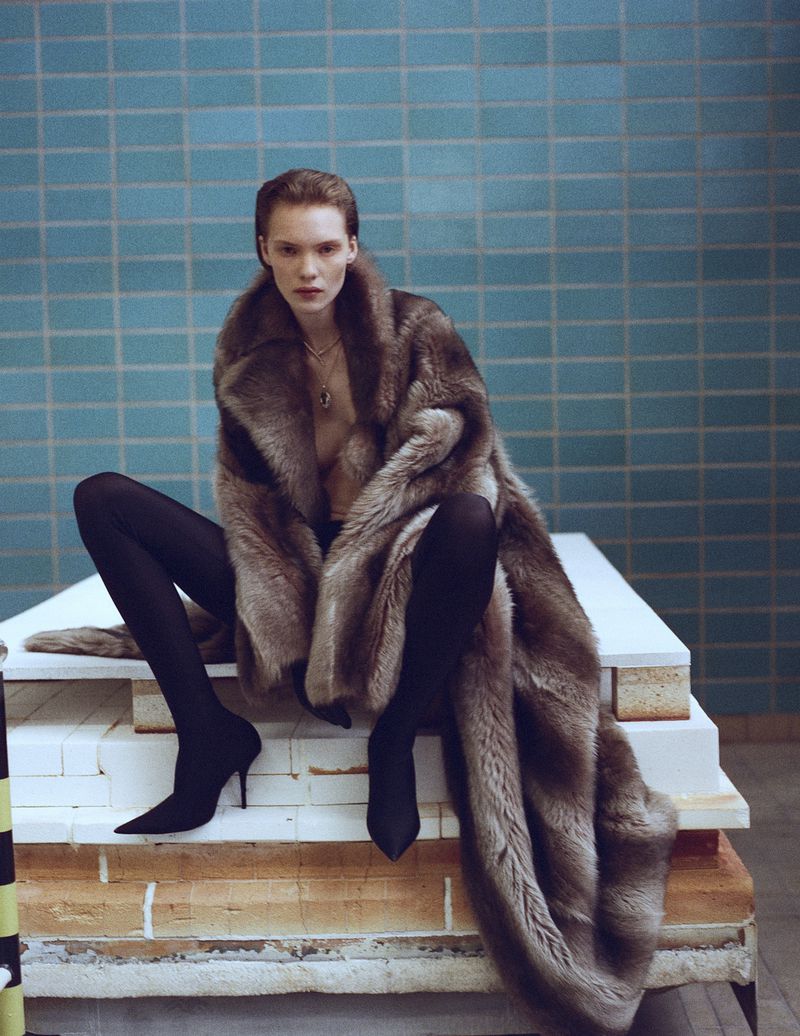 Penelope Ternes seated in an oversized brown fur coat, black tights, and pointed-toe stiletto boots against a blue tiled wall.