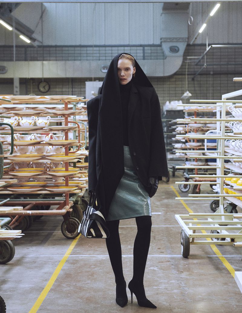 A fashion-forward woman in a black hooded coat and shiny green skirt poses in an industrial setting among rolling racks.