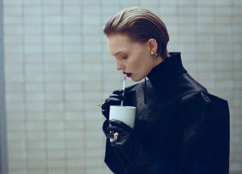 Blonde woman in a black top sips from a white cup through a straw. Red lipstick and black leather gloves add a dramatic touch.