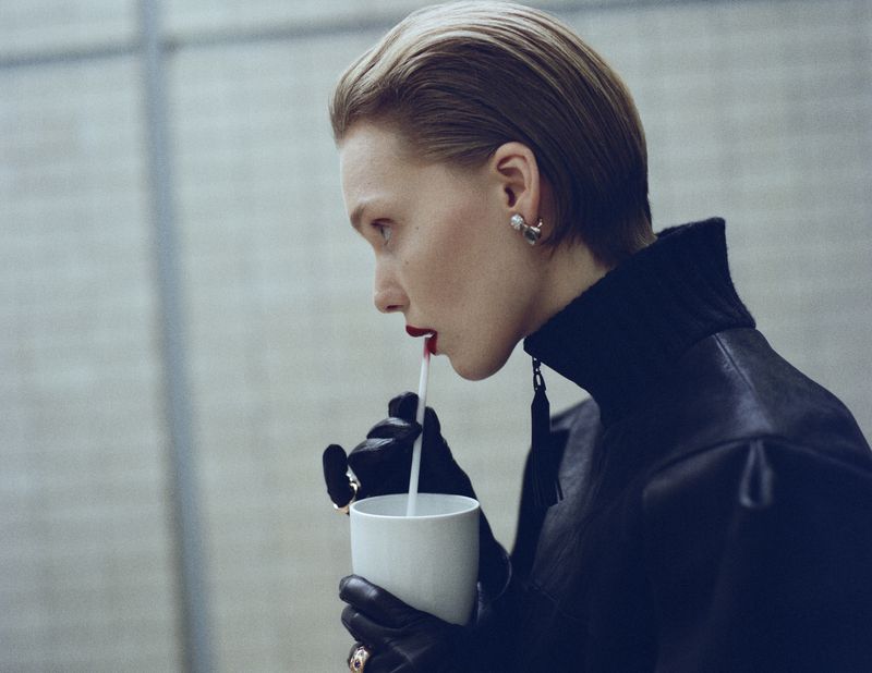 A light-skinned person with short hair drinks from a straw in an indoor setting, wearing a black jacket and dark gloves in a profile shot.