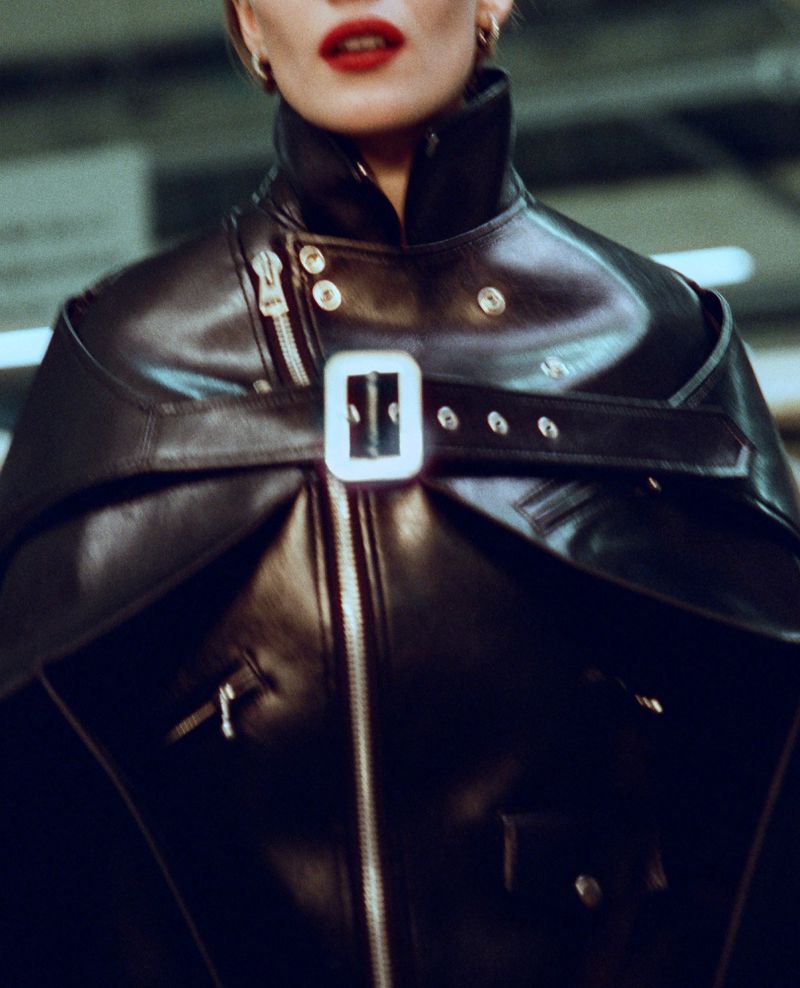 Close-up of a person in a black leather cape and jacket with silver hardware, featuring red lipstick and metallic earrings.