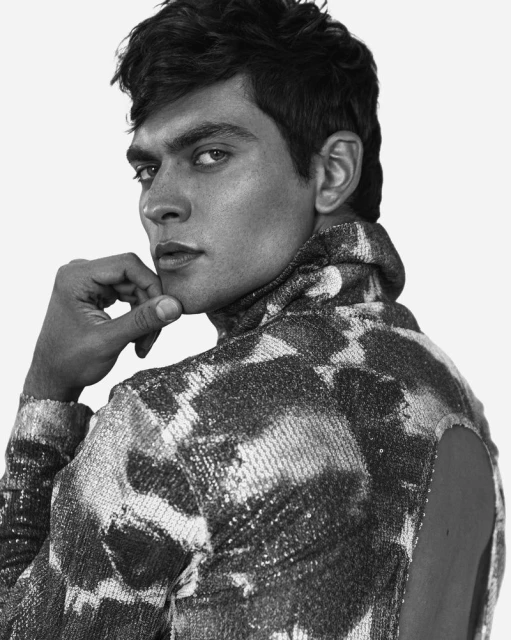 Monochrome portrait of a dark-haired male model in a patterned turtle neck sweater, hand to his chin, gazing over his shoulder.