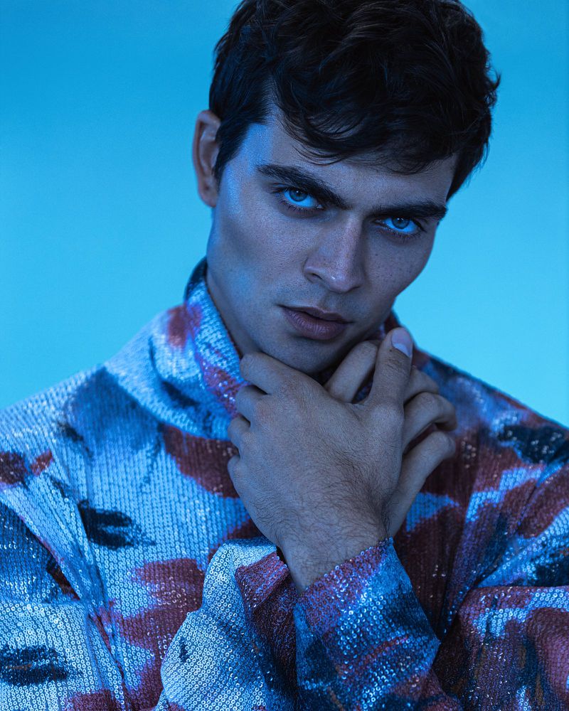 Close-up studio portrait of a young adult man with dark hair wearing a patterned jacket against a cool blue background.