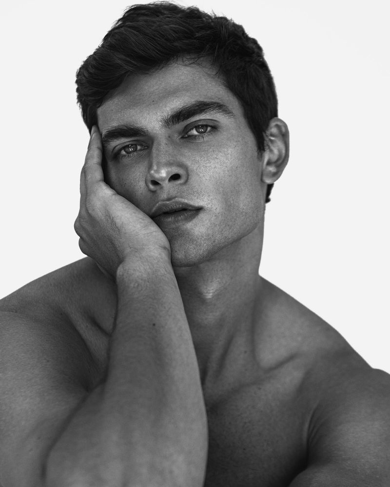 Portrait of a shirtless adult male with dark hair resting his head on his hand in front of a stark white backdrop in a photo studio.