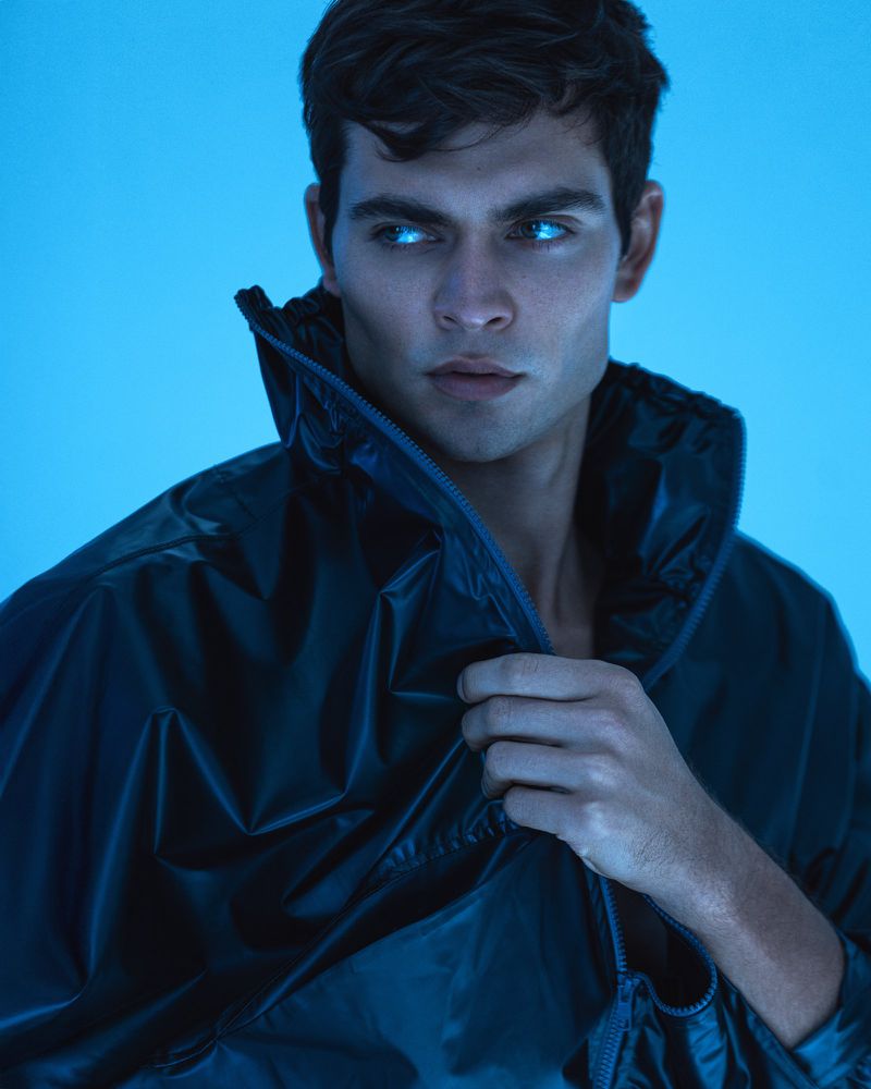Portrait of an adult male with dark hair wearing a black jacket, gazing off-camera in a studio environment with a blue color cast.