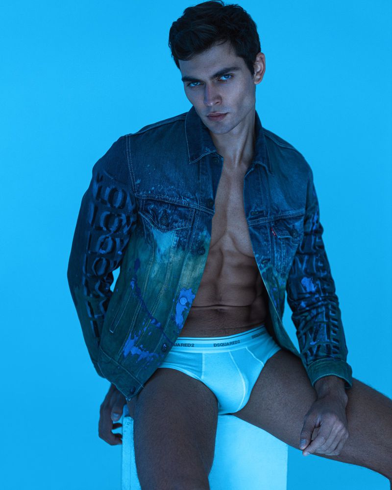 Portrait of a male model wearing a denim jacket and underwear against a vibrant blue backdrop in a fashion forward studio shot.