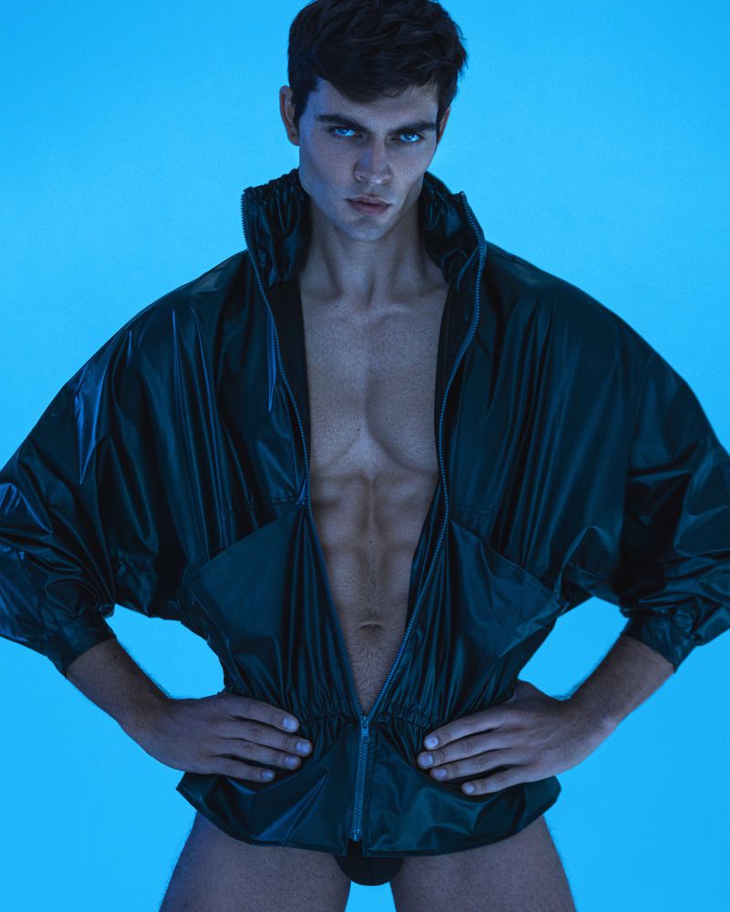 A muscular adult male poses in a studio with vibrant blue lighting wearing an unzipped shiny dark jacket and black briefs, staring intensely.