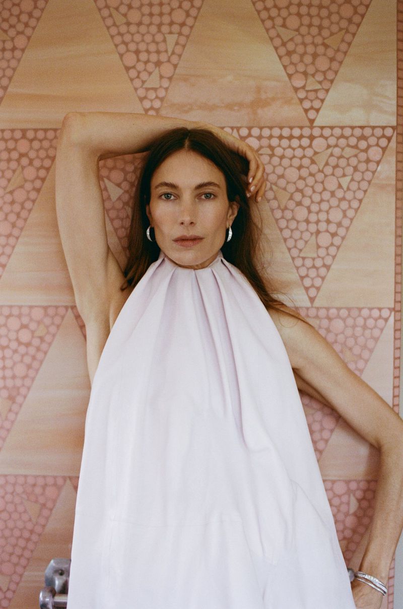 Model wearing a pleated white halter dress posing against a patterned geometric tiled wall