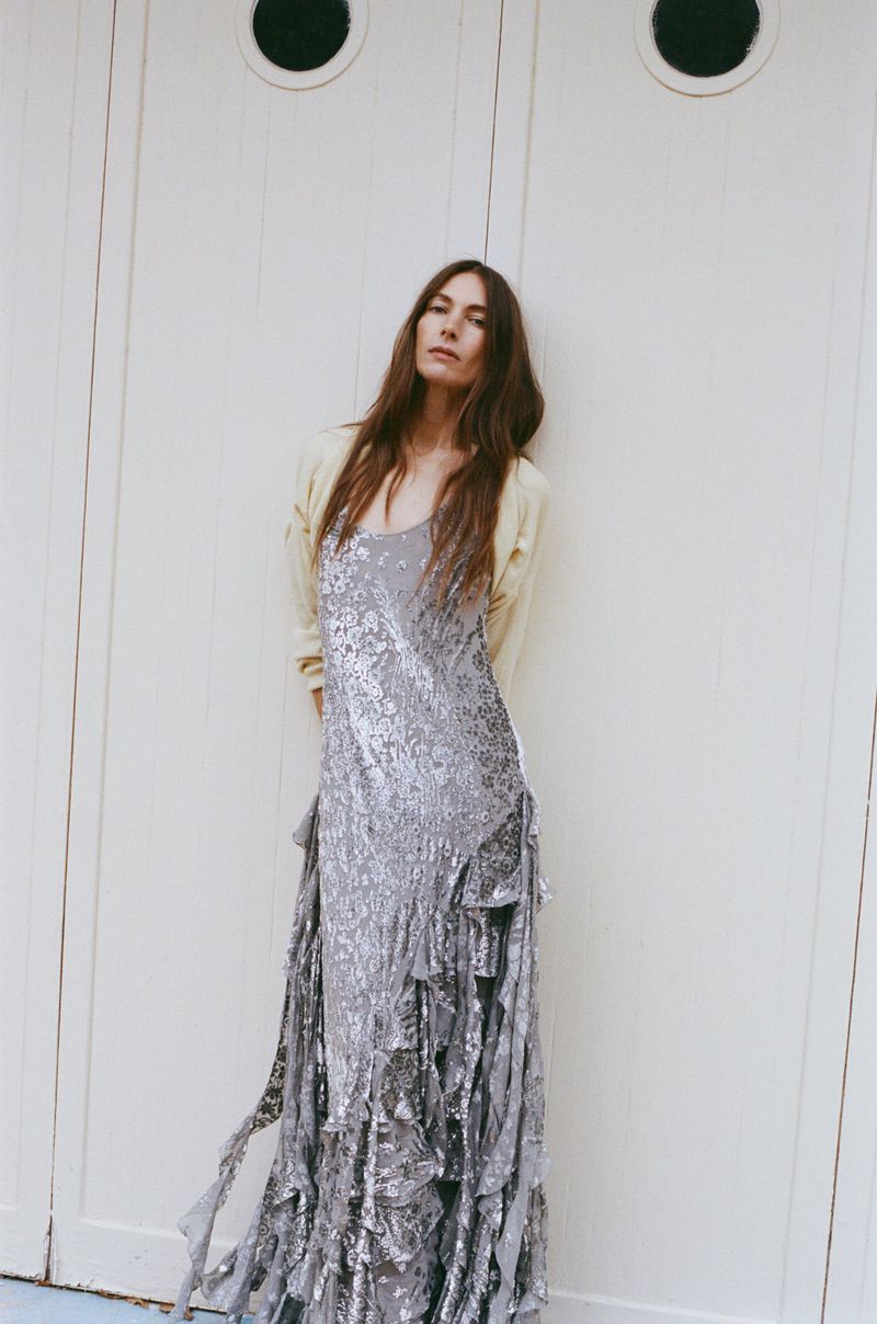 A tall woman with long brown hair poses in front of white doors, wearing a light yellow cardigan and a floor-length silver sequined dress.