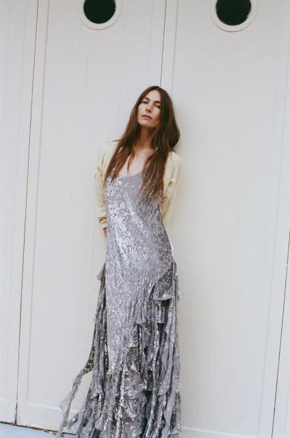 A tall woman with long brown hair poses in front of white doors, wearing a light yellow cardigan and a floor-length silver sequined dress.
