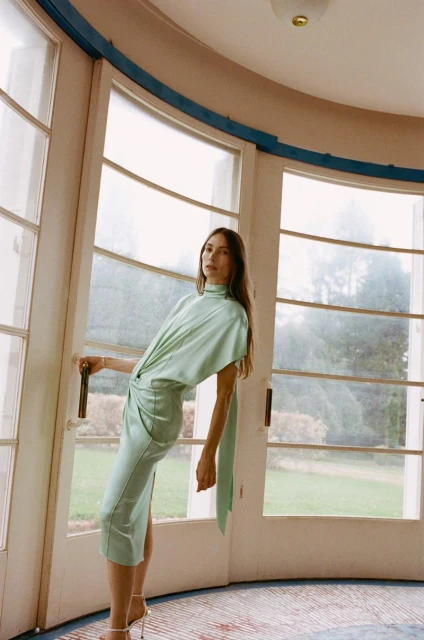 Adult female wearing a light green dress poses near a large window in an indoor space with natural lighting and tiled floors.