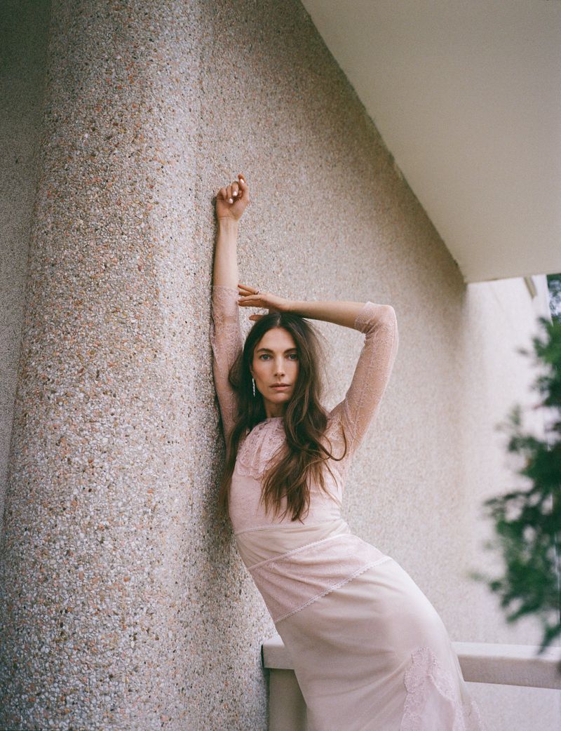 A slender adult woman with long brown hair leans against a speckled concrete wall wearing a light pink, full-sleeved dress, outdoors.
