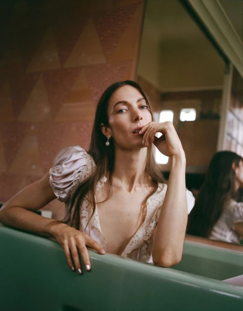 Adult female with long brown hair in a silk and lace dress poses in a green bathtub against a background with pink geometric wall tiles.