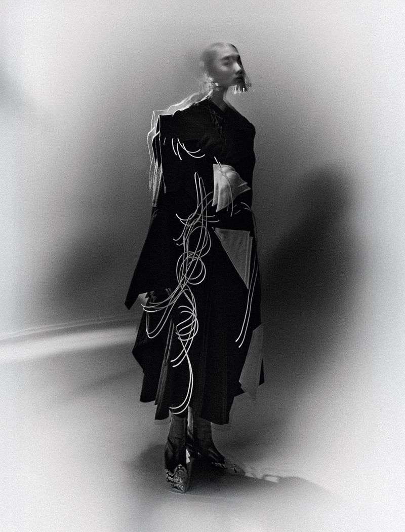 Monochrome fashion shot of an adult female wearing a dark coat with light-colored abstract design, statement earrings, and heeled shoes in a studio.