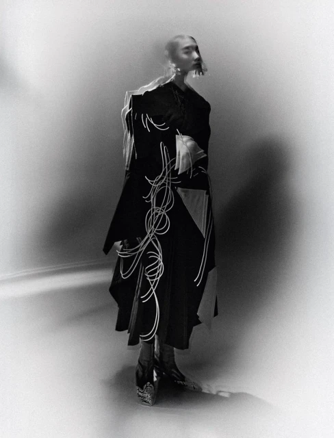 Monochrome fashion shot of an adult female wearing a dark coat with light-colored abstract design, statement earrings, and heeled shoes in a studio.