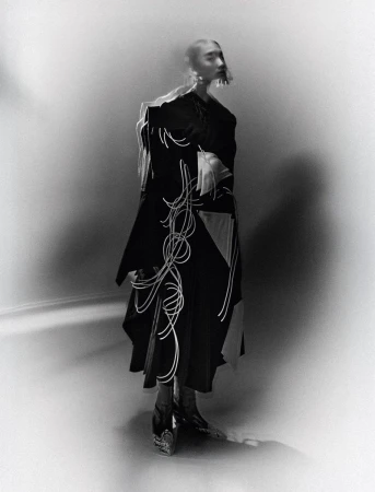 Monochrome fashion shot of an adult female wearing a dark coat with light-colored abstract design, statement earrings, and heeled shoes in a studio.