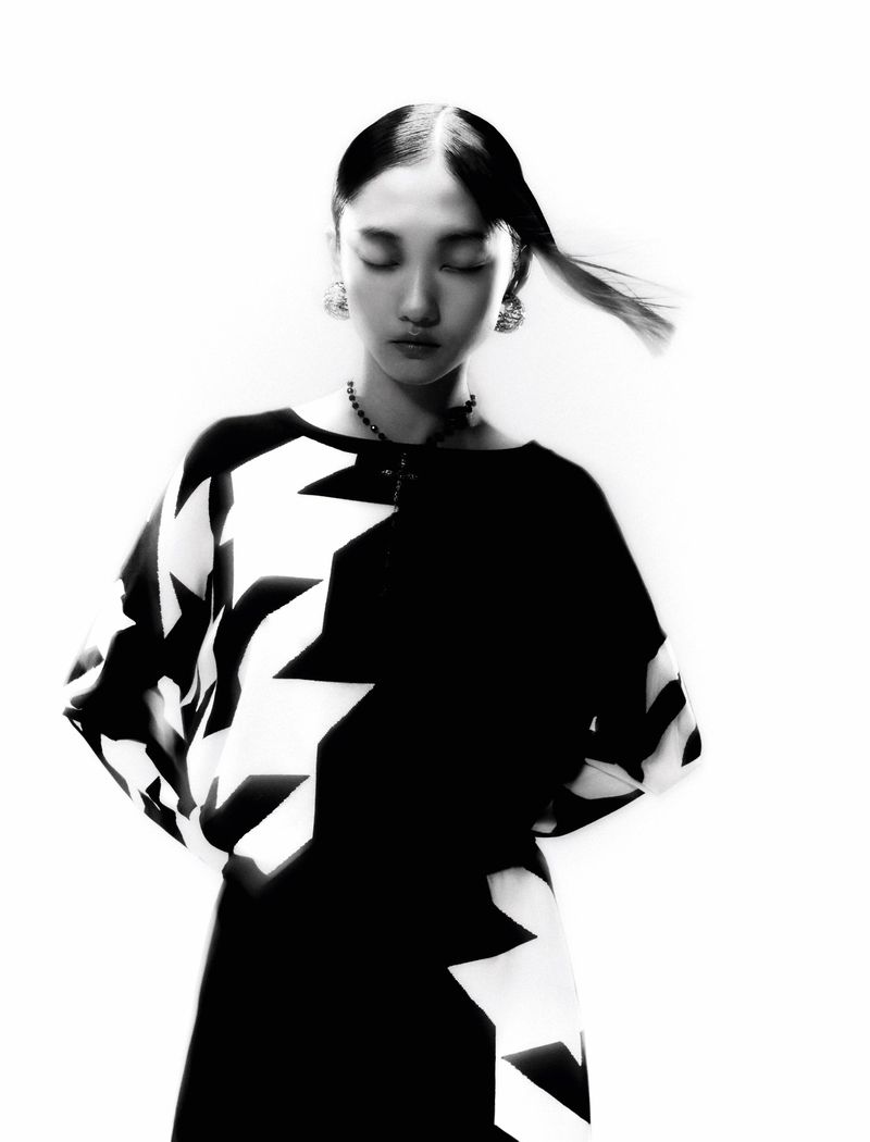 Black and white portrait of an Asian woman with closed eyes and a geometric-patterned dress against a bright, neutral background.