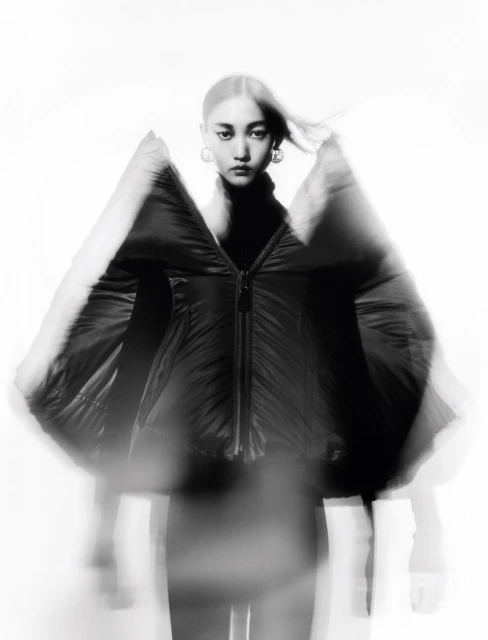 Asian woman in black puffer jacket poses against a white background with motion blur effect for fashion or beauty editorial content.