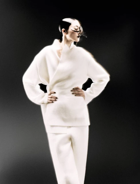 Fashion shot featuring a woman with dramatic eye makeup and a structured white outfit against a dark background in a studio setting.