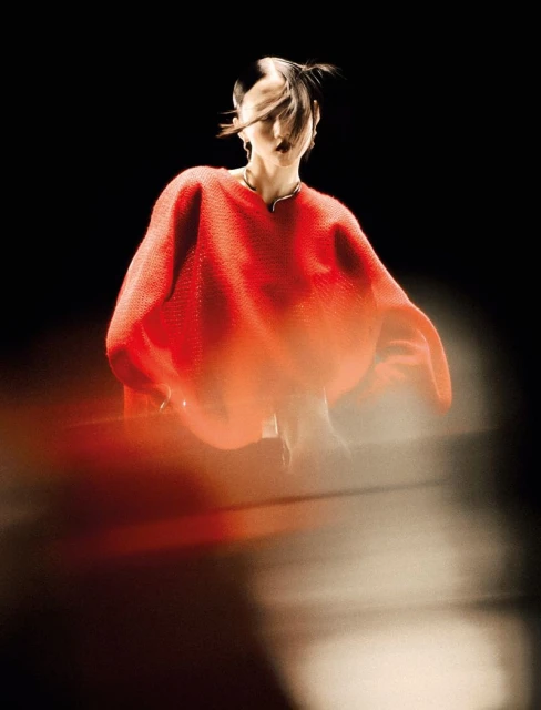 Stylish woman in a red textured top poses against a dark background, with long exposure creating a blurred effect.