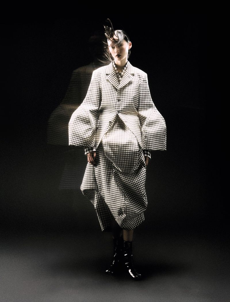 A woman poses in a black and white checkered coat and skirt in a dimly lit studio with a blurred figure behind her.