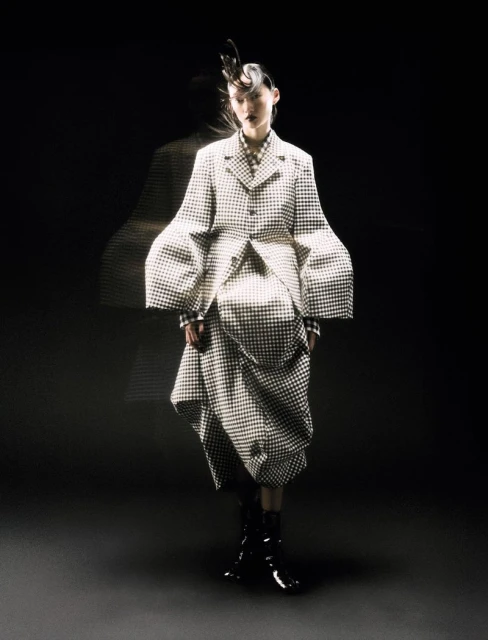 A woman poses in a black and white checkered coat and skirt in a dimly lit studio with a blurred figure behind her.