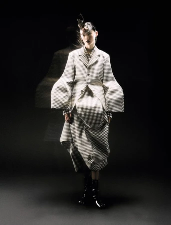 A woman poses in a black and white checkered coat and skirt in a dimly lit studio with a blurred figure behind her.