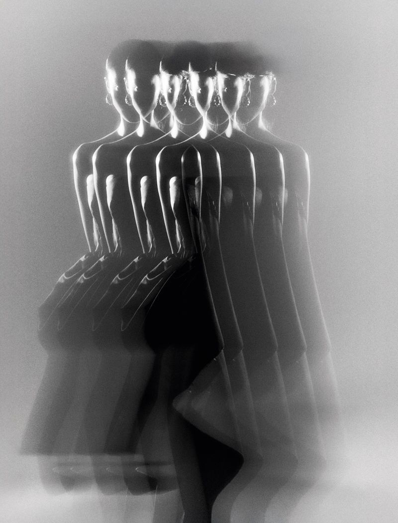 Monochromatic long exposure shot of a woman in a black dress and jewelry, her figure multiplied and blurred in motion against a white background.