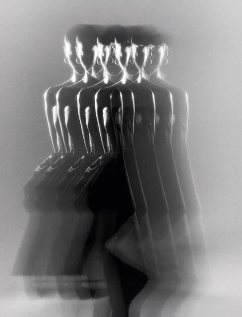 Monochromatic long exposure shot of a woman in a black dress and jewelry, her figure multiplied and blurred in motion against a white background.