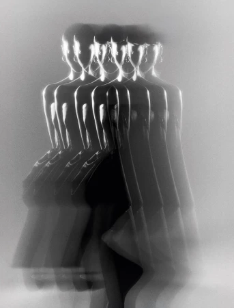 Monochromatic long exposure shot of a woman in a black dress and jewelry, her figure multiplied and blurred in motion against a white background.