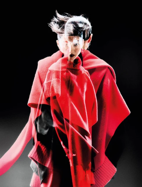 A woman with a windblown hairstyle and dark lipstick wears a red sweater against a black background in a fashion portrait.