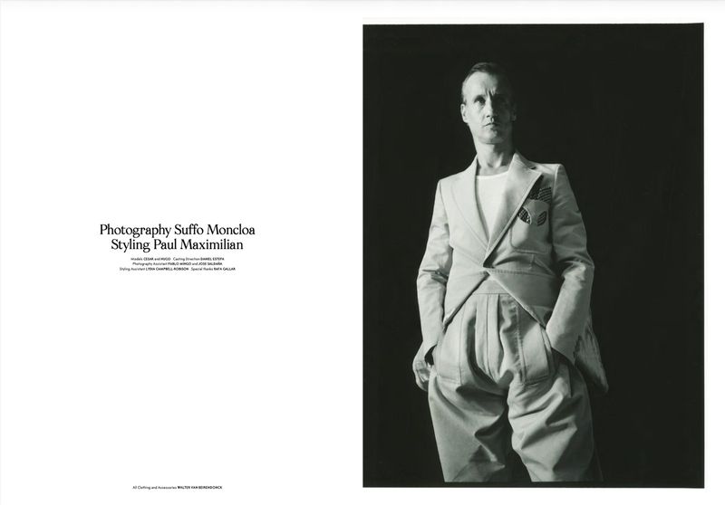 Black and white fashion portrait of a slim adult male wearing a suit jacket, t-shirt, and wide-legged trousers against a dark backdrop.