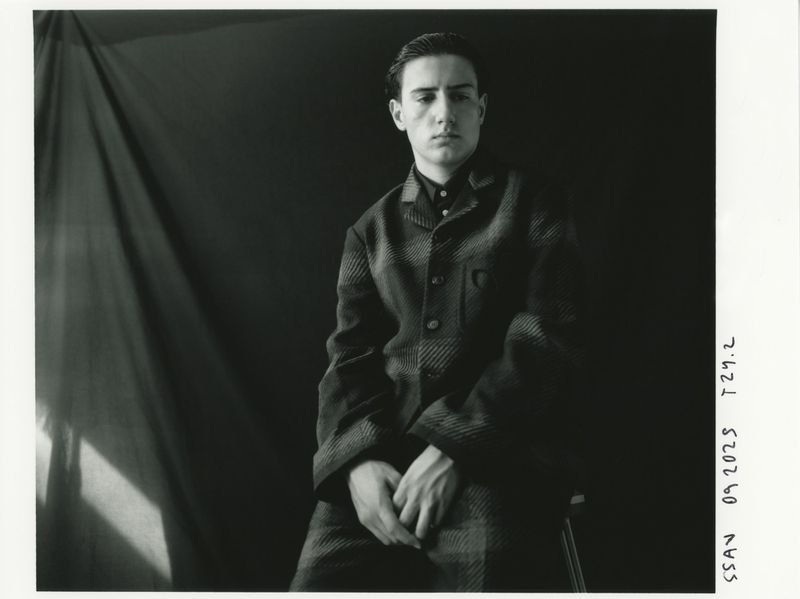 Monochrome portrait of a young adult male in a patterned suit, seated against a dark backdrop, bathed in soft studio light.