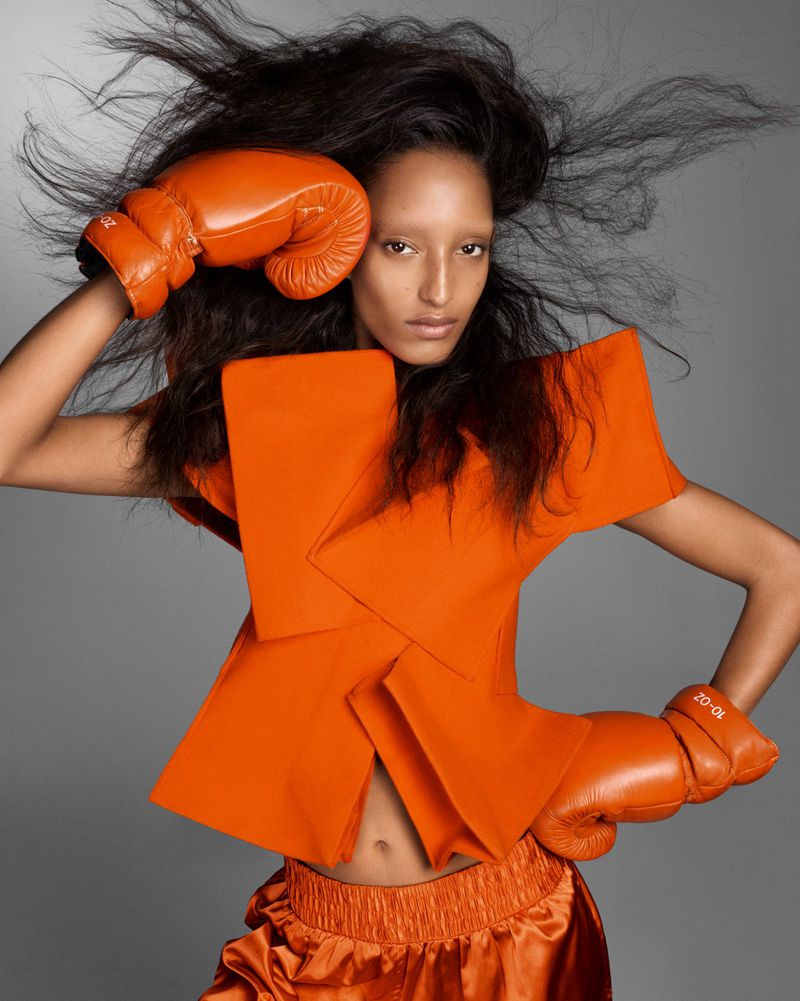 Fashion shot of a woman with orange boxing gloves and an avant-garde top, set against a grey backdrop in a studio setting.