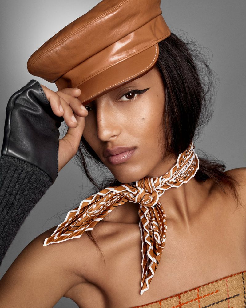 Close-up studio shot of a woman in a brown leather cap and patterned scarf, looking directly at the camera against a grey background.