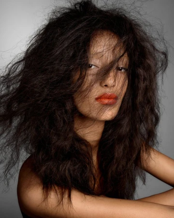 Close-up portrait of model Mona Tougaard with voluminous dark hair and bright orange lipstick against a grey background