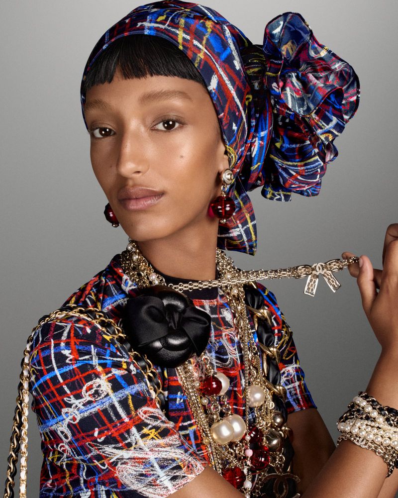 Adult female with headscarf and layered jewelry, including beaded necklaces and earrings, poses in a studio with soft, neutral lighting.